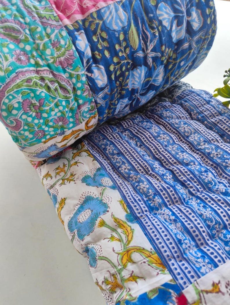 May include: A quilted cushion featuring a patchwork design with various floral and paisley patterns in shades of blue, white, pink, and green. The fabric is stitched with a grid pattern, showcasing multiple distinct textile sections joined together.