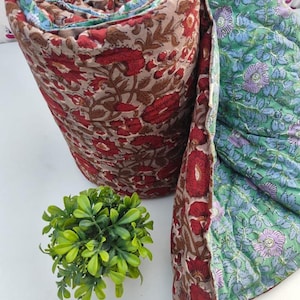 May include: A rolled quilted blanket featuring a floral pattern with red and brown tones on one side and a contrasting green background with purple and blue floral motifs on the reverse. The fabric displays intricate block-printed style details.