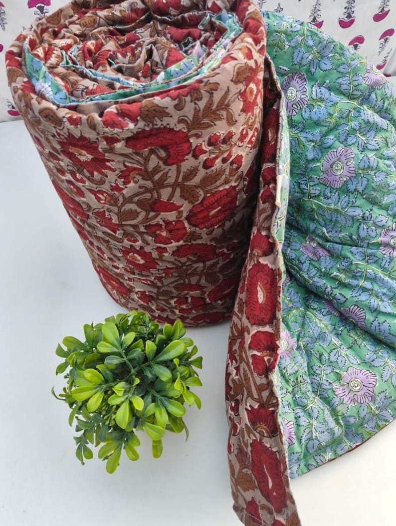 May include: A rolled quilted blanket featuring a floral pattern with red and brown tones on one side and a contrasting green background with purple and blue floral motifs on the reverse. The fabric displays intricate block-printed style details.