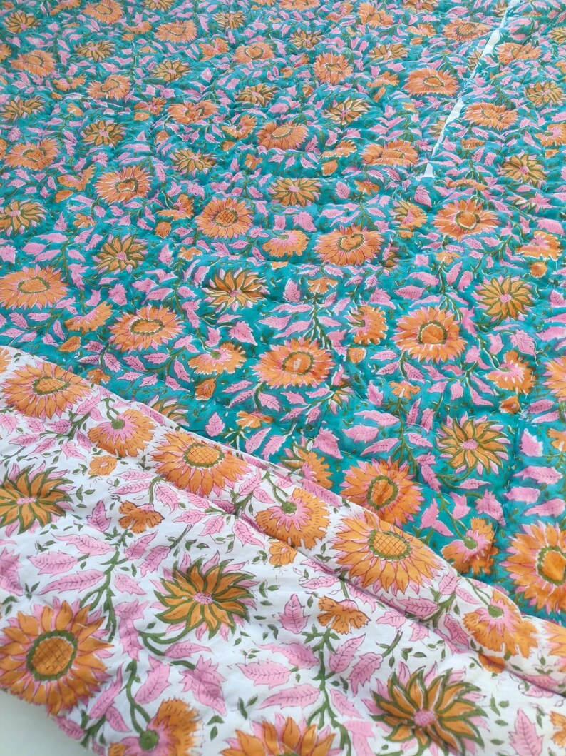 May include: A quilted blanket featuring a repeating floral pattern of orange sunflowers and pink leaves. One side has a vibrant teal background, while the folded corner reveals a white background with the same floral print and visible stitching.