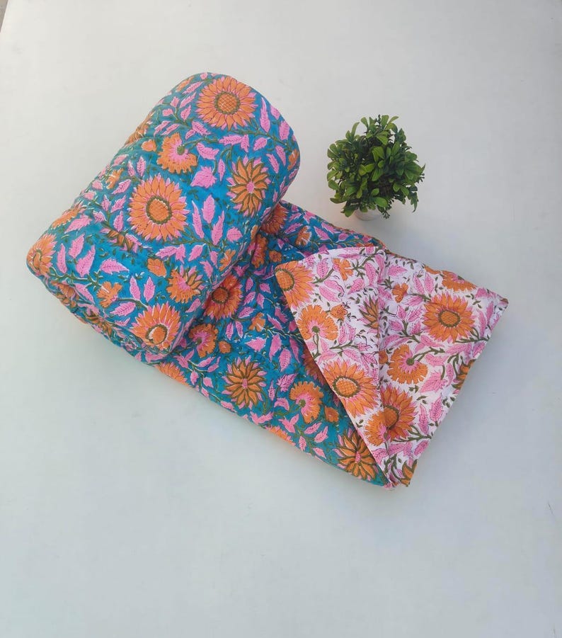 May include: A reversible quilted blanket featuring a vibrant floral pattern with orange sunflowers and pink leaves on a teal background, while the reverse side displays the same floral design in orange and pink on a white background.