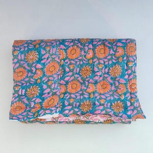 May include: A folded rectangular quilt featuring a vibrant teal background with a repeating pattern of orange sunflowers and pink leaves. The fabric displays visible quilted stitching lines throughout the surface, creating a textured appearance.