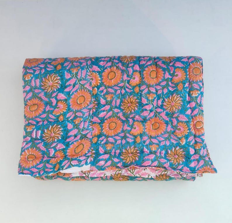 May include: A folded rectangular quilt featuring a vibrant teal background with a repeating pattern of orange sunflowers and pink leaves. The fabric displays visible quilted stitching lines throughout the surface, creating a textured appearance.