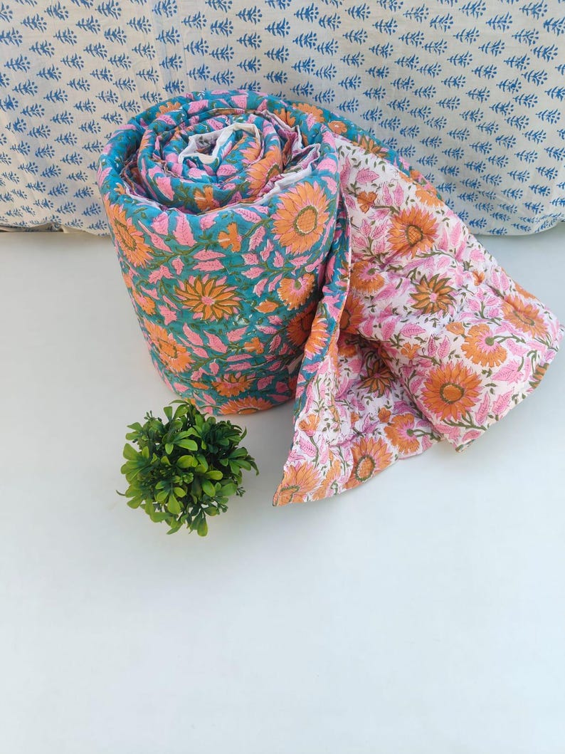 May include: A rolled cotton quilt featuring a vibrant floral pattern with orange sunflowers and pink leaves on a teal background, with a reverse side showing the same floral design in orange and pink on a white background.