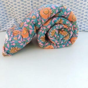 May include: A rolled cotton quilt featuring a vibrant teal background with a repeating floral pattern of orange sunflowers and pink leaves. The fabric has visible quilted stitching throughout, creating a soft, textured appearance.