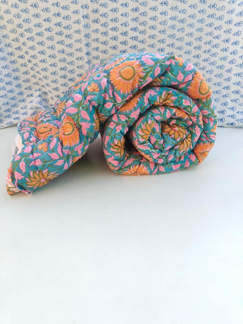 May include: A rolled cotton quilt featuring a vibrant teal background with a repeating floral pattern of orange sunflowers and pink leaves. The fabric has visible quilted stitching throughout, creating a soft, textured appearance.