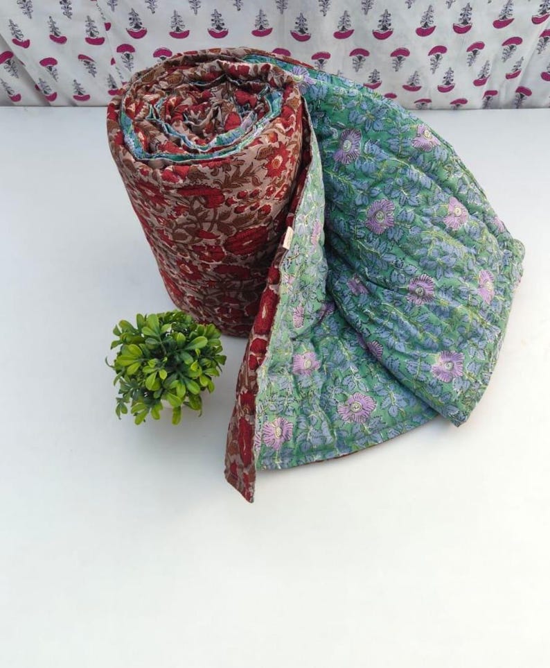 May include: A rolled quilted blanket featuring a deep red and brown floral block print on the exterior and a light green fabric with a purple and blue floral pattern on the interior, showcasing visible stitching throughout the textile surface.