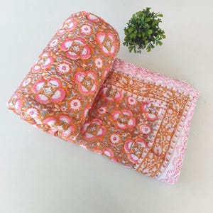 Puede incluir: Una colcha de algodón doblada con un vibrante estampado floral en tonos rosa brillante, naranja y blanco. El tejido muestra un diseño de estilo estampado en bloque con un borde decorativo, exhibiendo costuras intrincadas en toda la tela.
