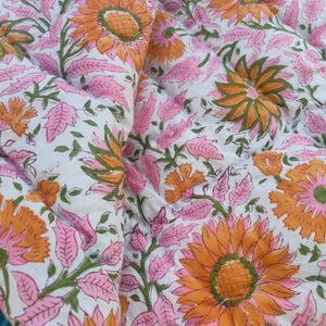 May include: A quilted fabric featuring a repeating floral pattern with large orange sunflowers, pink leaves, and green stems on a white background. The textile has visible stitching lines throughout, creating a textured, padded appearance.