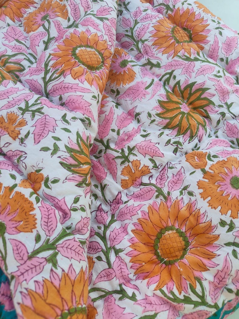 May include: A quilted fabric featuring a repeating floral pattern with large orange sunflowers, pink leaves, and green stems on a white background. The textile has visible stitching lines throughout, creating a textured, padded appearance.