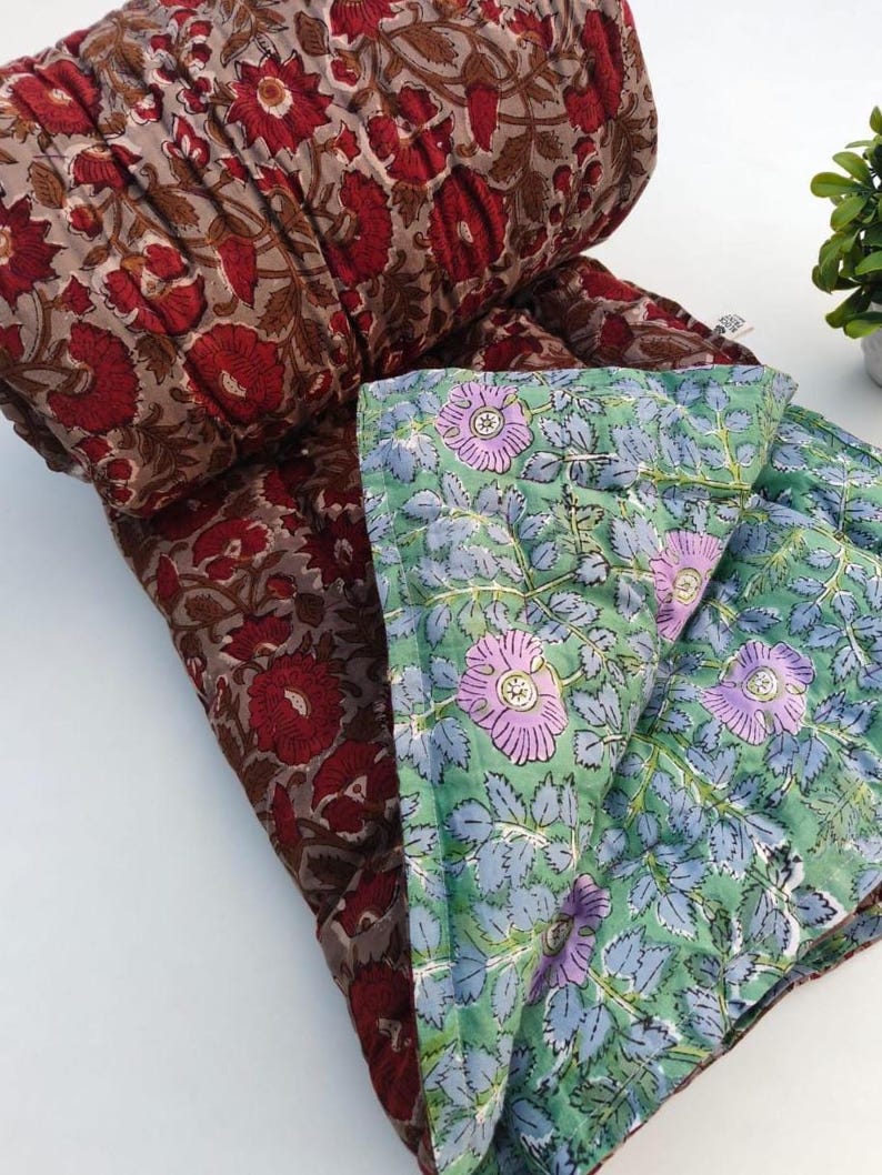 May include: A reversible quilted blanket featuring two distinct floral patterns. One side displays deep red flowers on a muted brown background, while the other side showcases light purple flowers with green leaves on a pale green background.