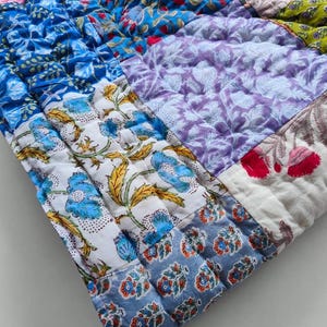 May include: A quilted textile featuring a patchwork of various floral and botanical patterns in vibrant shades of blue, purple, pink, green, and white. The fabric is stitched with a grid-like pattern, showcasing diverse block-printed designs.