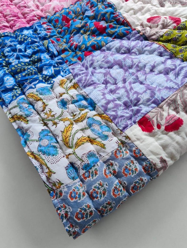 May include: A quilted textile featuring a patchwork of various floral and botanical patterns in vibrant shades of blue, purple, pink, green, and white. The fabric is stitched with a grid-like pattern, showcasing diverse block-printed designs.