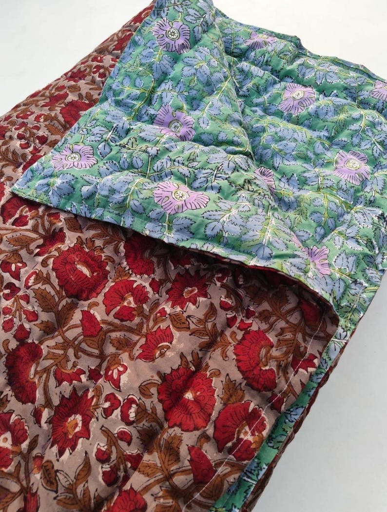 May include: A reversible quilted blanket featuring two distinct floral patterns. One side displays large red flowers with brown foliage on a light brown background, while the other side shows light purple flowers with green leaves on a light green background.