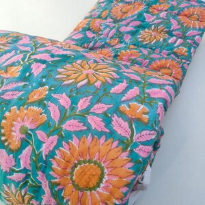 May include: A quilted textile featuring a vibrant teal background with a repeating floral pattern of large orange sunflowers and light pink leaves. The fabric has visible stitching lines throughout, creating a textured, layered appearance.