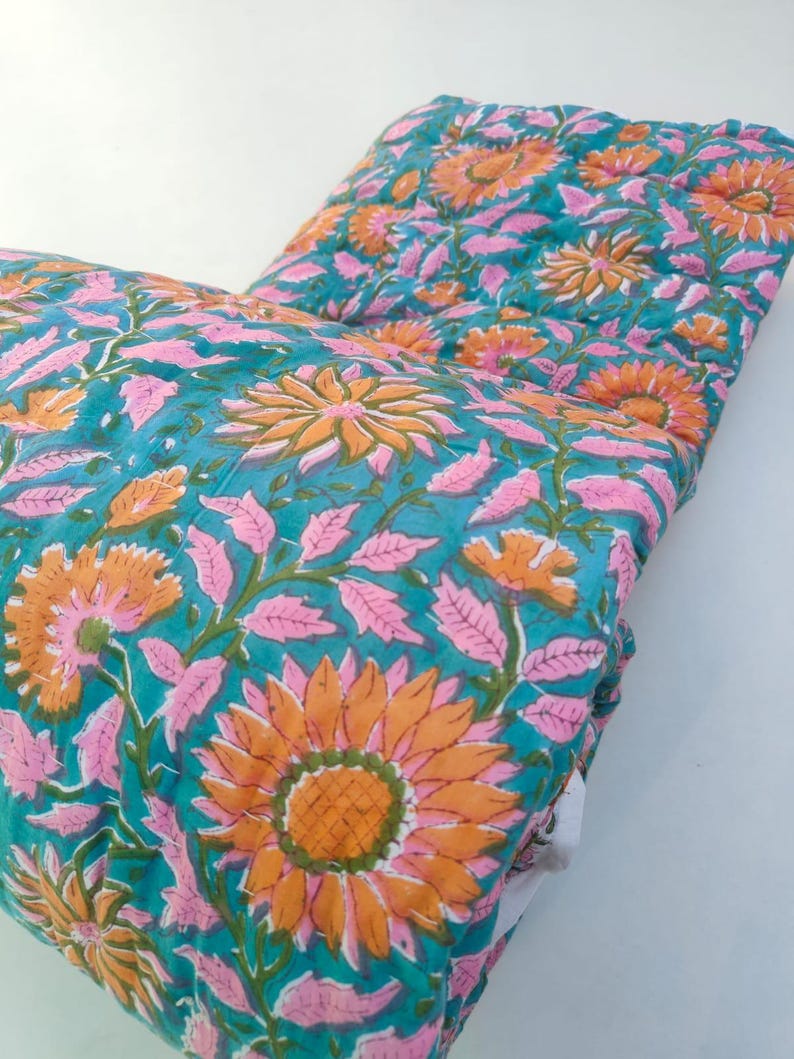 May include: A quilted textile featuring a vibrant teal background with a repeating floral pattern of large orange sunflowers and light pink leaves. The fabric has visible stitching lines throughout, creating a textured, layered appearance.