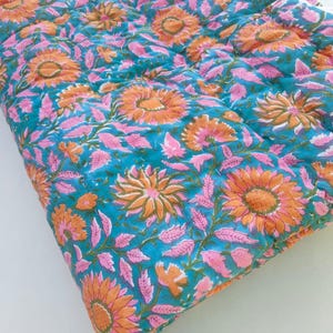 May include: A quilted textile featuring a vibrant teal background adorned with a repeating pattern of orange sunflowers and light pink leaves. The fabric displays visible stitching lines across the surface, creating a textured, layered appearance.