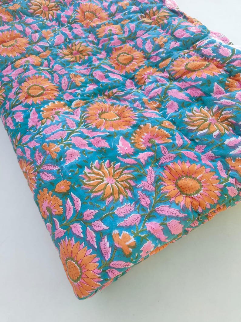 May include: A quilted textile featuring a vibrant teal background adorned with a repeating pattern of orange sunflowers and light pink leaves. The fabric displays visible stitching lines across the surface, creating a textured, layered appearance.