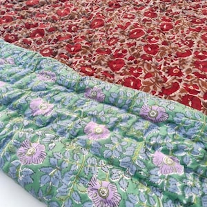 May include: A quilted textile featuring two distinct floral patterns. The top section displays a dense arrangement of red flowers on a tan background, while the folded bottom section shows light purple flowers on a light green background.