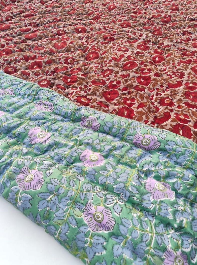 May include: A quilted textile featuring two distinct floral patterns. The top section displays a dense arrangement of red flowers on a tan background, while the folded bottom section shows light purple flowers on a light green background.