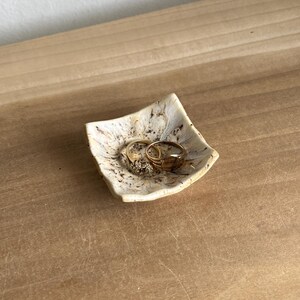 May include: A small, square, off-white ceramic trinket dish with a speckled brown and black pattern. The dish has slightly upturned, wavy edges and contains two gold-colored metal rings resting in the center of the basin.