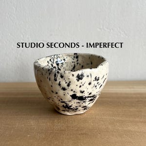 May include: A small, off-white ceramic bowl with irregular edges and a speckled black pattern. The text "STUDIO SECONDS - IMPERFECT" is above the bowl. The bowl is on a wooden surface.