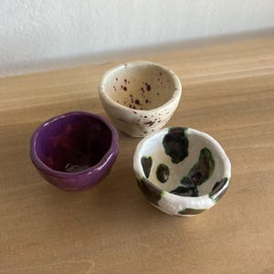 May include: Three small ceramic bowls in a close-up shot. One is deep purple, another is speckled beige, and the third has a white base with black and green patterns. The bowls are on a wooden surface.