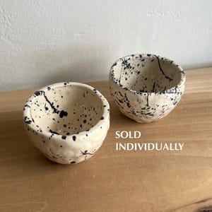 May include: Two small, round ceramic bowls with an off-white base color and a black speckled paint splatter pattern. The bowls have a textured, organic shape. The text "SOLD INDIVIDUALLY" is printed in white on the right side of the image.