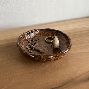 May include: A small, round, brown ceramic trinket dish with a speckled, glossy glaze. Inside the dish rests a delicate gold-toned chain necklace with a small pendant and a gold-toned ring featuring a smooth, cream-colored stone inlay.