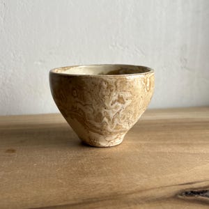 May include: A small, round ceramic bowl featuring a marbled pattern of tan, beige, and cream tones. The bowl has a smooth, glossy finish with a slightly uneven, organic rim and a tapered base, showcasing a blend of earthy, neutral colors throughout.