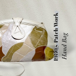 May include: A 1980's Patch Work Hand Bag featuring a multi-colored, textured, crocodile-embossed leather design in shades of cream, olive green, and brown. The bag has a curved top, a central decorative leather knot detail, and a thin strap.