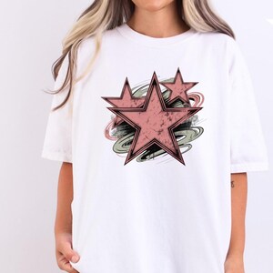 May include: White t-shirt featuring a distressed design of three pink stars with black outlines, set against a backdrop of green, gray, and black brushstrokes. The shirt is a comfortable, casual style.