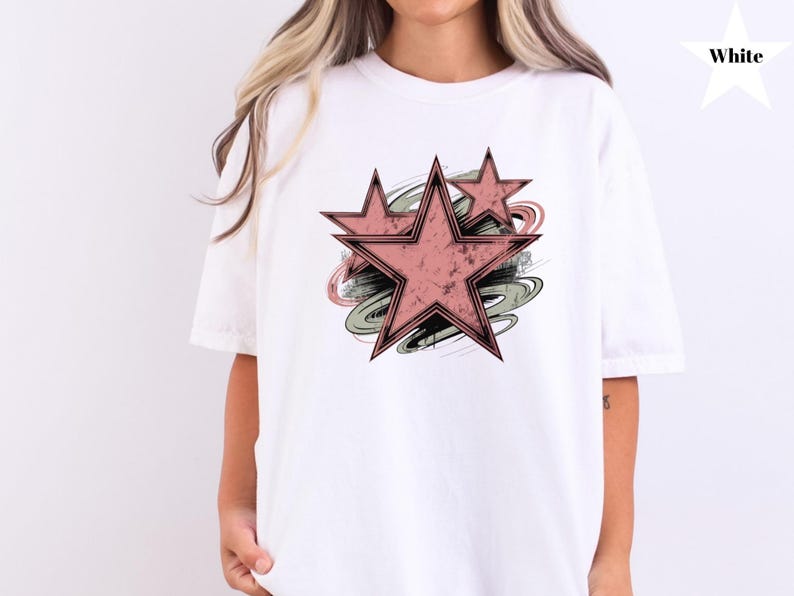 May include: White t-shirt featuring a distressed design of three pink stars with black outlines, set against a backdrop of green, gray, and black brushstrokes. The shirt is a comfortable, casual style.