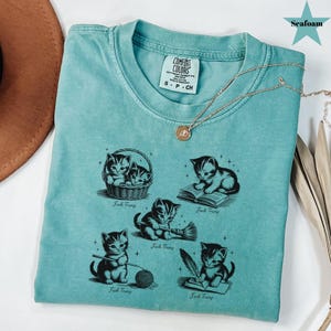 May include: Seafoam green t-shirt with a graphic of kittens in various poses, including reading and writing. The text "Fuck Trump" is below each kitten illustration. A gold necklace with a pendant is draped across the shirt.