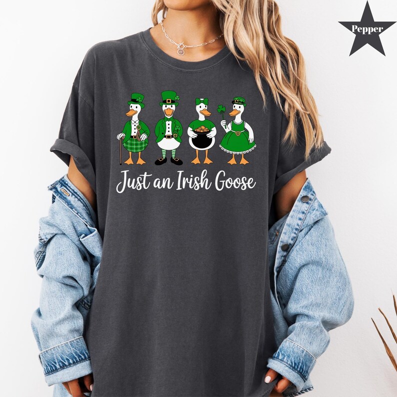 Puede incluir: Camiseta gris oscuro con un gr&aacute;fico de cuatro gansos vestidos con atuendos irlandeses. Los gansos llevan sombreros, chalecos y vestidos verdes, sosteniendo un tr&eacute;bol, una olla de oro y un bast&oacute;n. El texto "Just an Irish Goose" est&aacute; impreso debajo.