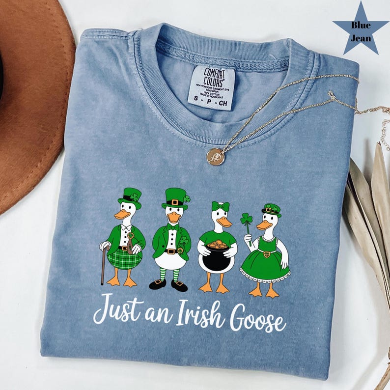Puede incluir: Camiseta azul vaquero con un gr&aacute;fico de cuatro gansos de dibujos animados vestidos de verde para el D&iacute;a de San Patricio. Los gansos sostienen un bast&oacute;n, una olla de oro y un tr&eacute;bol de cuatro hojas. El texto "Just an Irish Goose" est&aacute; impreso debajo.