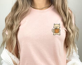 May include: A light pink t-shirt featuring a cartoon cat holding an orange pumpkin. The cat is smiling, and the design is in a simple, outlined style. The shirt is made of a soft material and is suitable for casual wear.