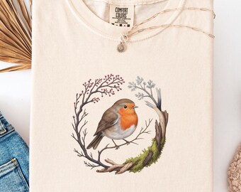 May include: Ivory-colored t-shirt featuring a watercolor illustration of a robin perched on a branch within a circular wreath of twigs and moss. The shirt has a Comfort Colors tag and a gold necklace with a pendant. The word "Ivory" is displayed in a star shape.