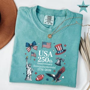 May include: Seafoam green t-shirt with a patriotic design celebrating the USA's 250th anniversary. The design includes the Statue of Liberty, American flags, a top hat, a football, and an eagle. The text reads "USA 250th Anniversary Semiquincentennial 1776-2026".
