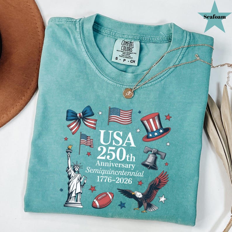 May include: Seafoam green t-shirt with a patriotic design celebrating the USA's 250th anniversary. The design includes the Statue of Liberty, American flags, a top hat, a football, and an eagle. The text reads "USA 250th Anniversary Semiquincentennial 1776-2026".