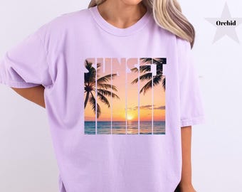 May include: A light purple t-shirt with a sunset graphic. The graphic features palm trees, ocean, and a vibrant sunset with the word "SUNSET" in white. The shirt is a comfortable, oversized fit.