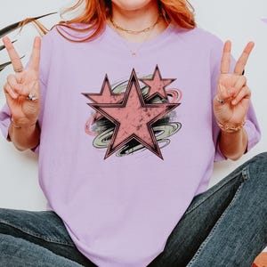 May include: A light purple t-shirt featuring a distressed pink star design with black outlines and a swirl background. The word "Orchid" is in the upper right corner. The model is wearing blue jeans.