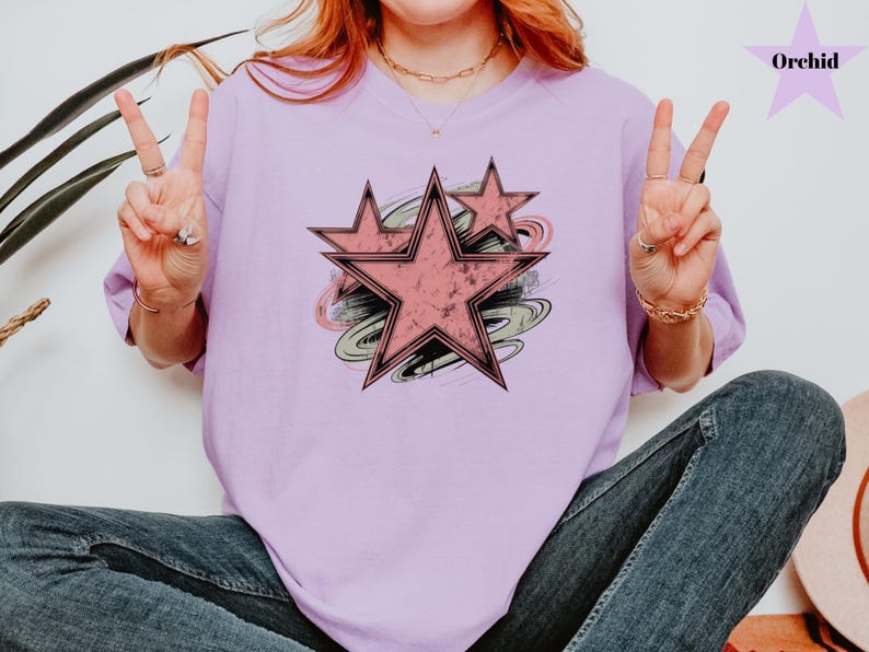 May include: A light purple t-shirt featuring a distressed pink star design with black outlines and a swirl background. The word "Orchid" is in the upper right corner. The model is wearing blue jeans.