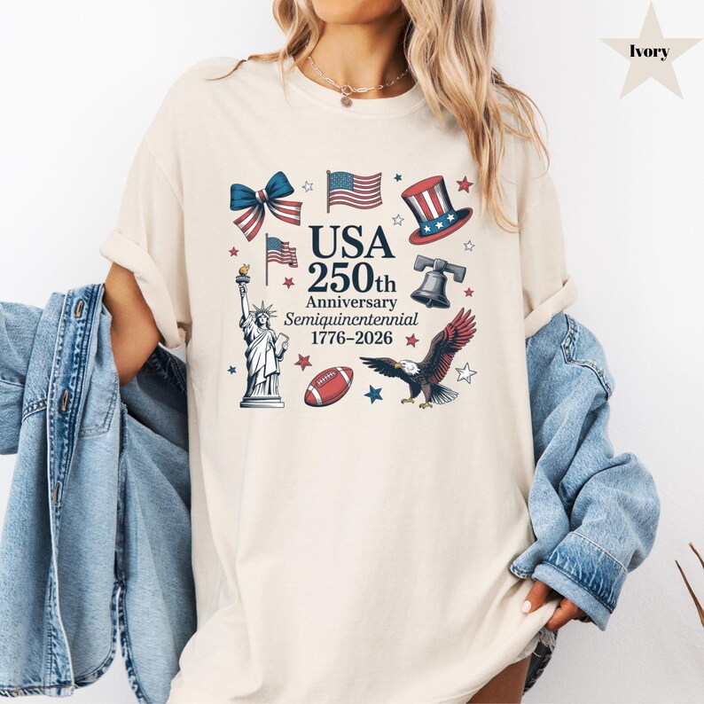 May include: Ivory-colored t-shirt featuring a patriotic design celebrating the USA's 250th anniversary. The graphic includes the Statue of Liberty, an American flag, a football, an eagle, and the text "USA 250th Anniversary Semiquincentennial 1776-2026."
