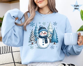 May include: Light blue sweatshirt featuring a snowman with a blue and black plaid hat and scarf, surrounded by pine trees and snowflakes. A small blue bird is perched on a branch. The sweatshirt is a solid light blue color.