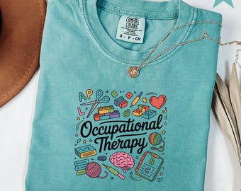May include: Seafoam green t-shirt with the words "Occupational Therapy" and colorful illustrations of scissors, blocks, a heart, and a brain. A gold necklace is draped across the shirt.