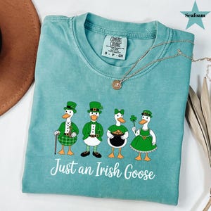 Puede incluir: Camiseta verde mar con un gr&aacute;fico de cuatro gansos de dibujos animados vestidos para el D&iacute;a de San Patricio. Los gansos llevan sombreros, chaquetas y vestidos verdes, y sostienen objetos como una olla de oro y un tr&eacute;bol. El texto "Just an Irish Goose" est&aacute; debajo.