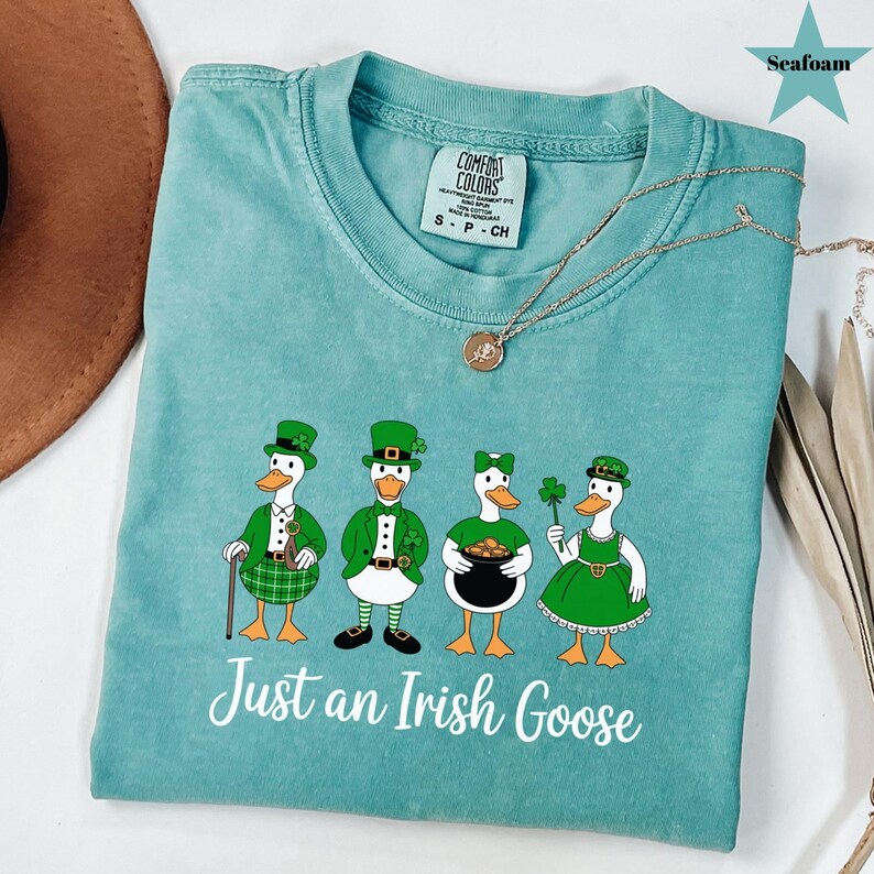 Puede incluir: Camiseta verde mar con un gr&aacute;fico de cuatro gansos de dibujos animados vestidos para el D&iacute;a de San Patricio. Los gansos llevan sombreros, chaquetas y vestidos verdes, y sostienen objetos como una olla de oro y un tr&eacute;bol. El texto "Just an Irish Goose" est&aacute; debajo.