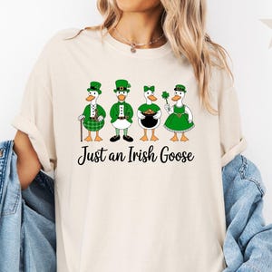 Puede incluir: Camiseta color marfil con un gr&aacute;fico de cuatro gansos vestidos con atuendos del D&iacute;a de San Patricio. Cada ganso lleva un sombrero y un atuendo verdes, uno con un tr&eacute;bol y otro con una olla de oro. El texto "Just an Irish Goose" est&aacute; debajo.