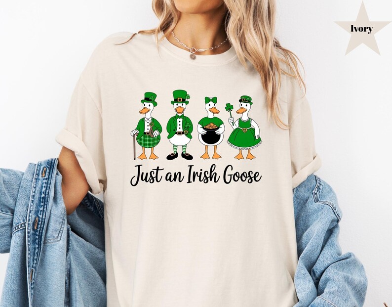 Puede incluir: Camiseta color marfil con un gr&aacute;fico de cuatro gansos vestidos con atuendos del D&iacute;a de San Patricio. Cada ganso lleva un sombrero y un atuendo verdes, uno con un tr&eacute;bol y otro con una olla de oro. El texto "Just an Irish Goose" est&aacute; debajo.